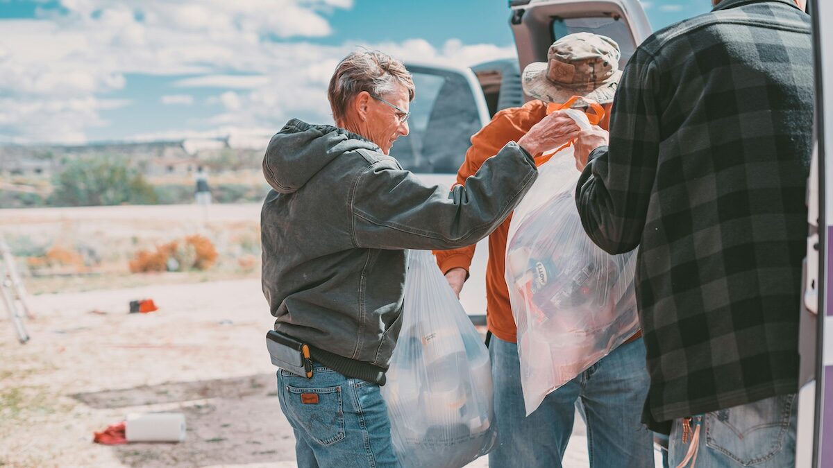 Three people stand by a van, holding large clear plastic bags, appearing to load or unload items outdoors on a sunny day.