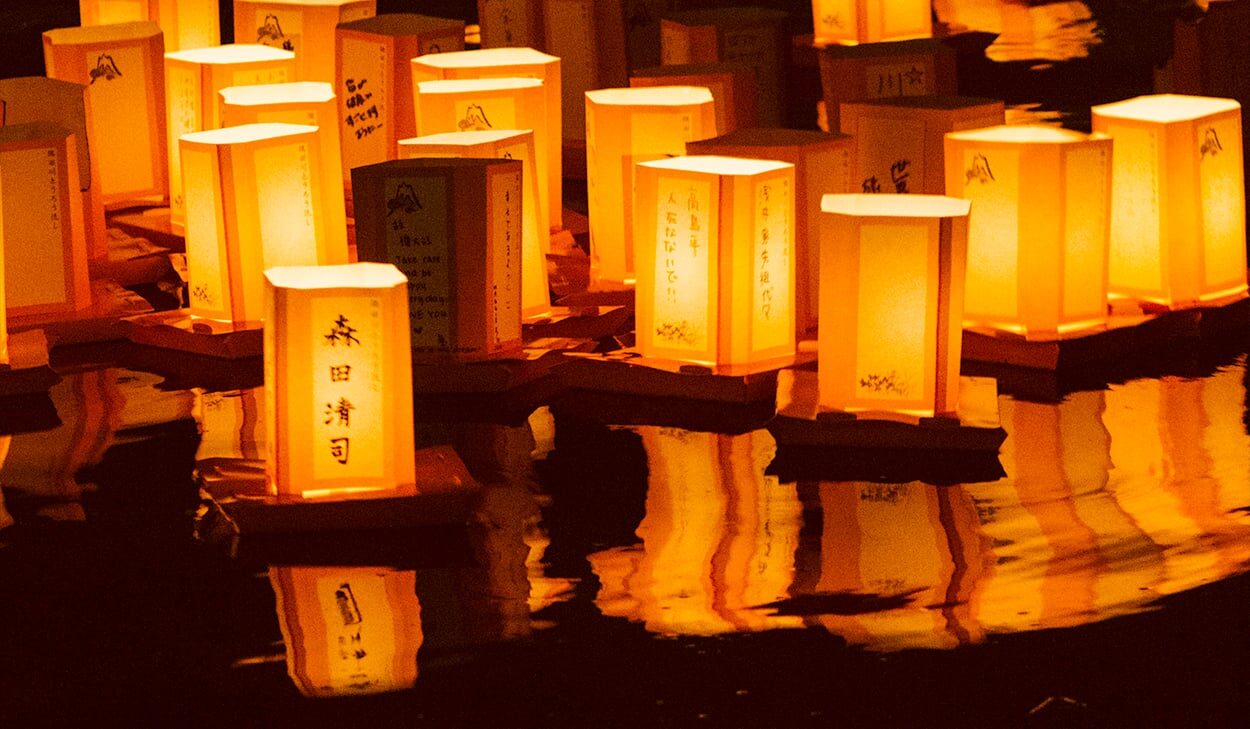 Paper lanterns with Japanese writing float on water at night, casting a warm orange glow and reflections on the surface.