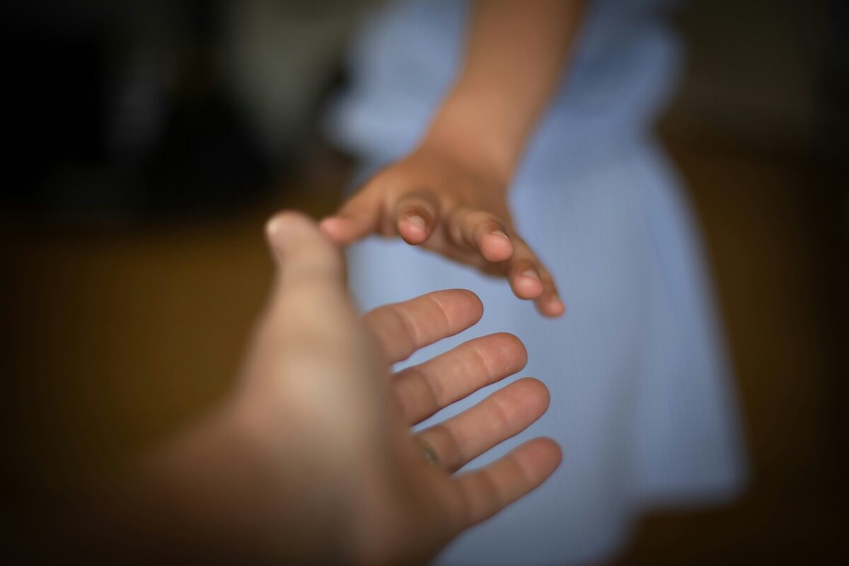 A close-up of two hands reaching towards each other, one belonging to a child in a light blue outfit and the other to an adult, against a blurred background.