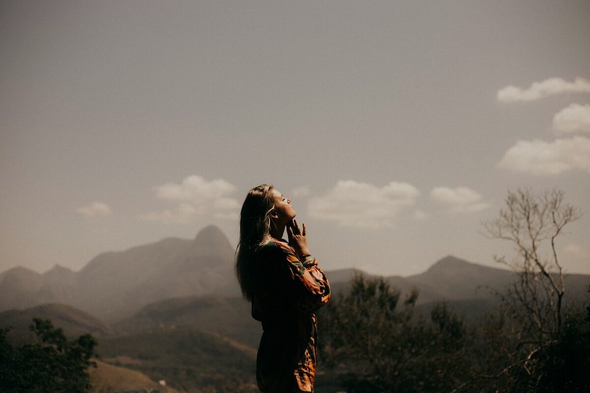 A person in a long-sleeved outfit stands outdoors, gazing at distant mountains under a partly cloudy sky during daylight.