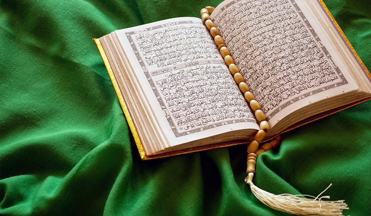 An open Quran with a wooden prayer bead strand rests on a green cloth.