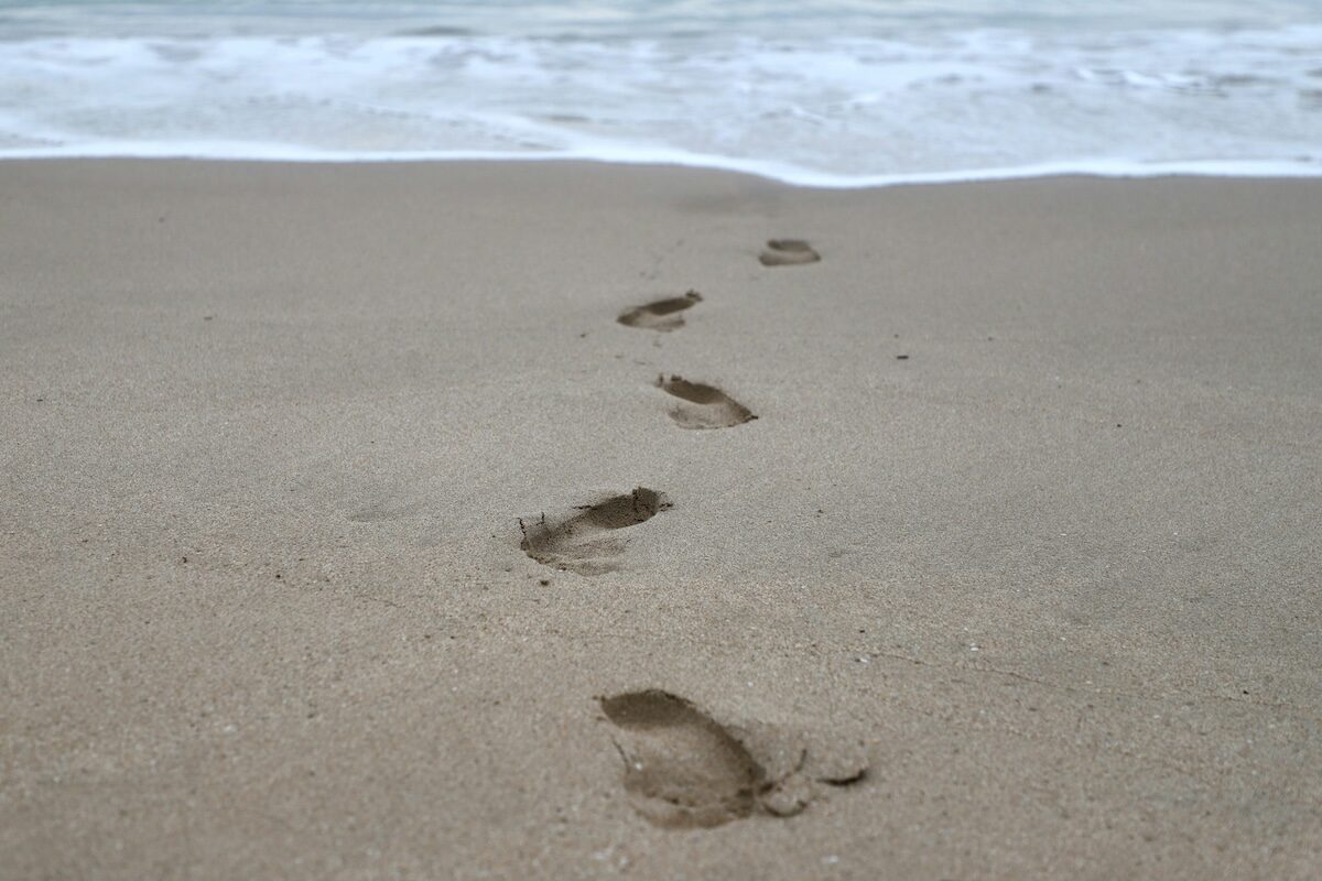 A series of human footprints in wet sand lead toward the shoreline, with gentle waves approaching in the background.