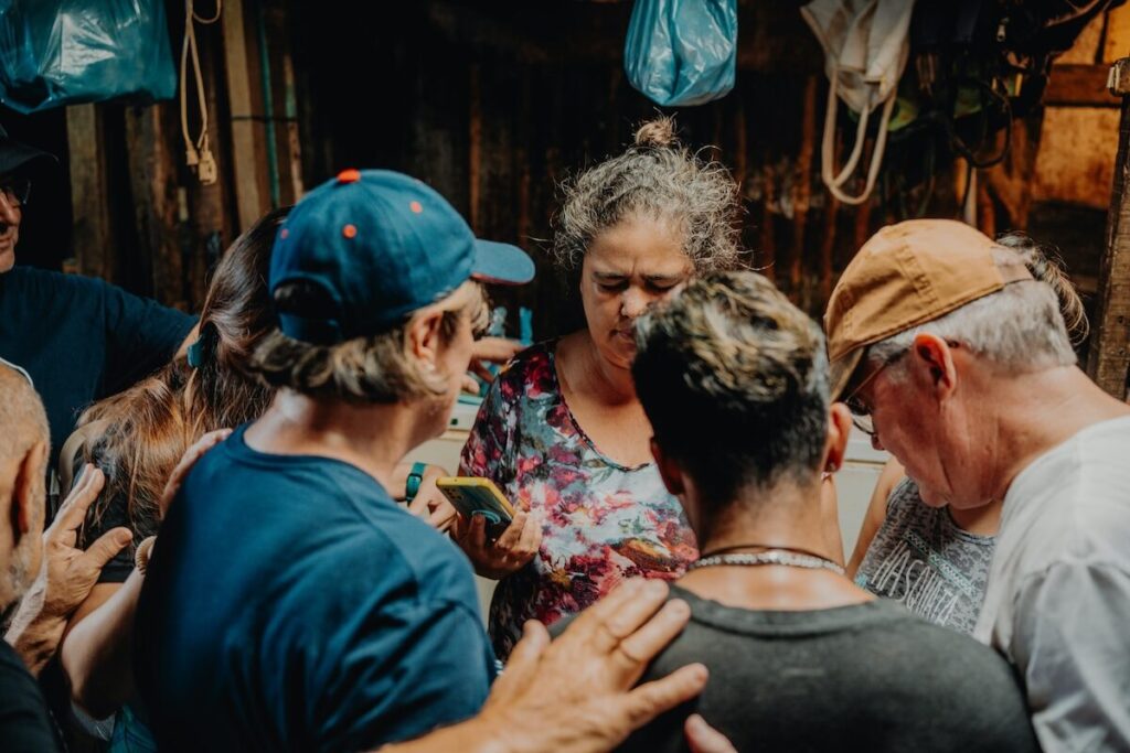 A group of people stand close together in a circle indoors, some with hands on each others shoulders, appearing engaged in a shared moment or activity.