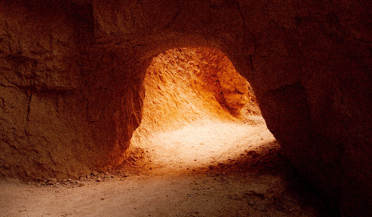 A narrow dirt path curves through a reddish-brown cave tunnel, illuminated by natural light from an opening ahead.