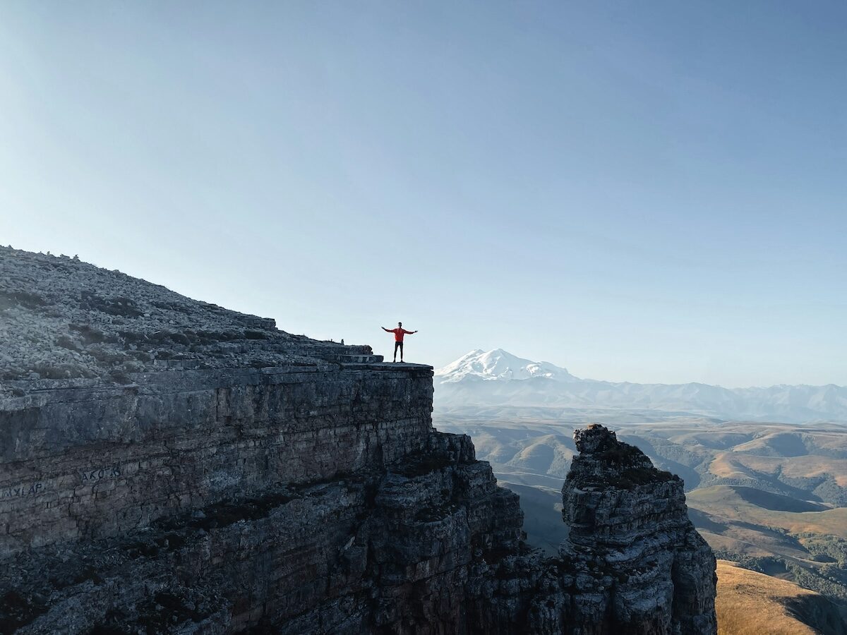 A person in a red jacket stands with arms outstretched on the edge of a rocky cliff, with mountains visible in the background under a clear sky.