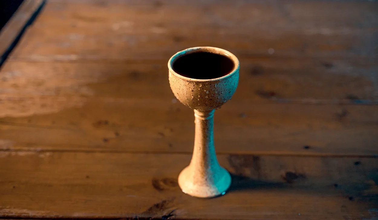 A handmade ceramic chalice filled with a dark liquid sits on a wooden table.