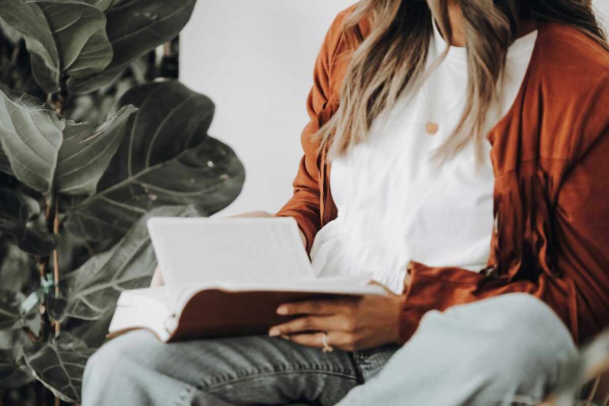 A person wearing a white shirt and brown sweater sits next to a large plant, holding and reading an open book.