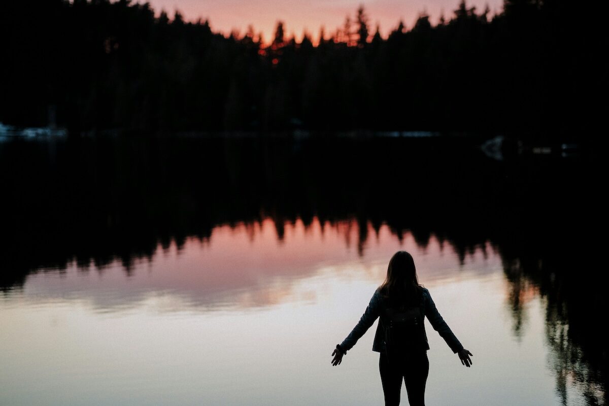 Silhouette of a person standing by a calm lake at sunset, with trees and a colorful sky reflected in the water.