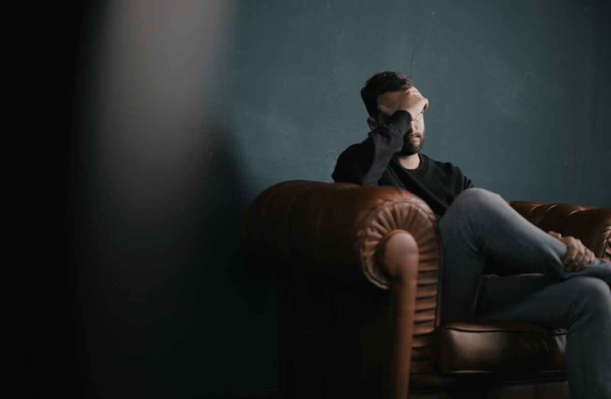 A man sits on a brown leather couch with his legs crossed, resting his head on his hand and looking distressed against a dark background.