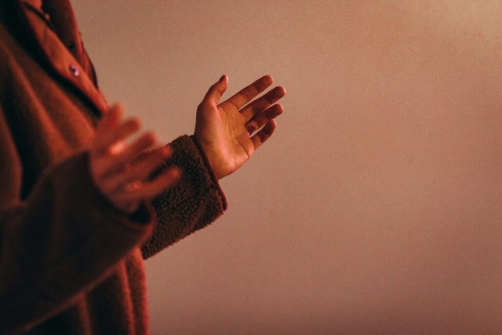 A person wearing a brown jacket holds their hands up, palms open, against a plain, softly lit background.