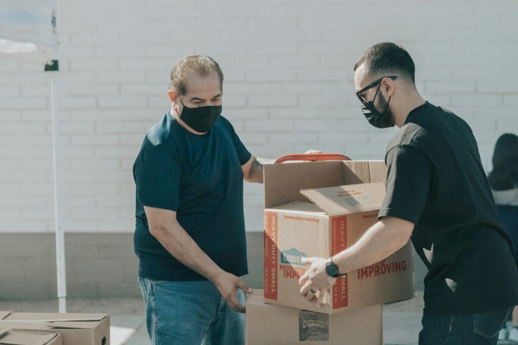 Two men wearing face masks handle cardboard boxes outdoors against a white brick wall.