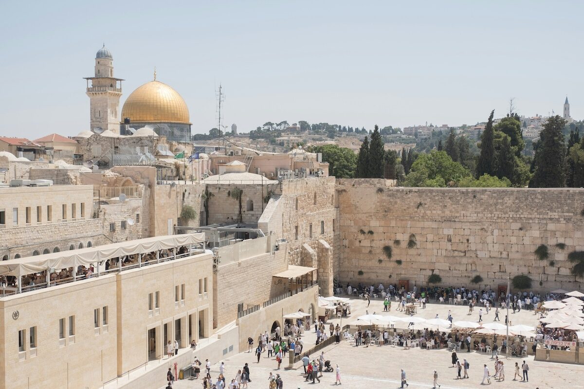 View of the Western Wall plaza in Jerusalem, with the Dome of the Rock and trees visible in the background under a clear sky.