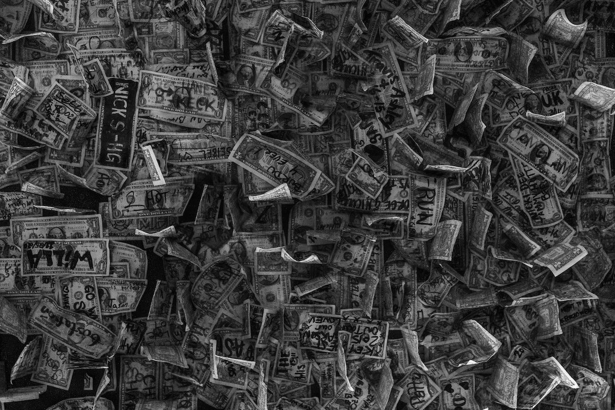 A dense collage of dollar bills, many crumpled and overlapping, some with handwritten messages and drawings. Image is in black and white.