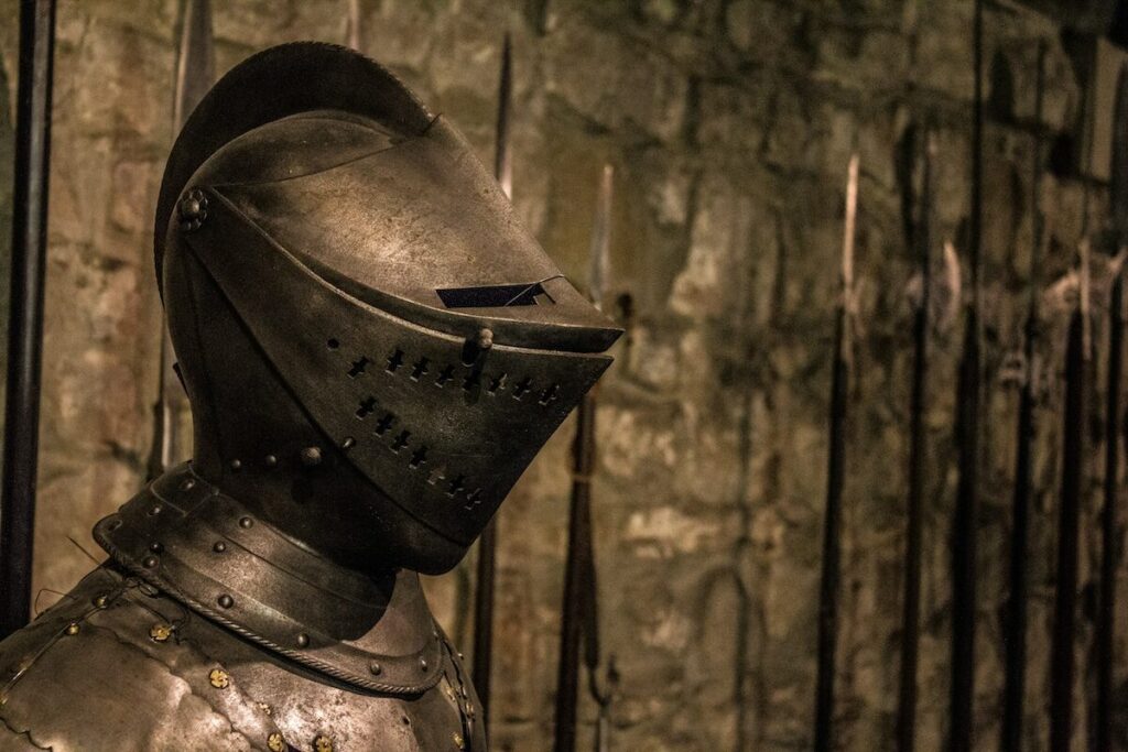A medieval suit of armor with a closed helmet is displayed in front of a stone wall and several upright spears.