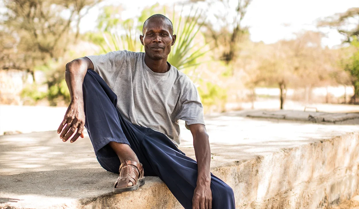 A man wearing a gray t-shirt, blue pants, and brown shoes sits on a concrete ledge outdoors with trees and sunlight in the background.
