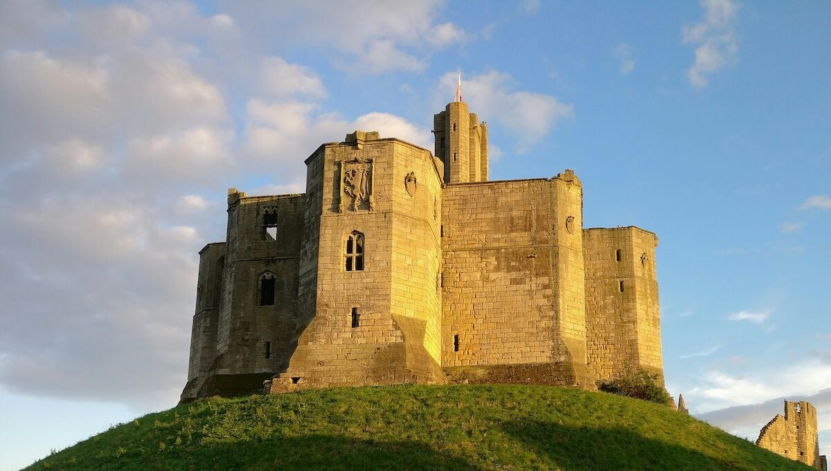 Stone castle with round towers and central keep, situated on a grassy hill, shown under a cloudy sky with sunlight illuminating the structure.