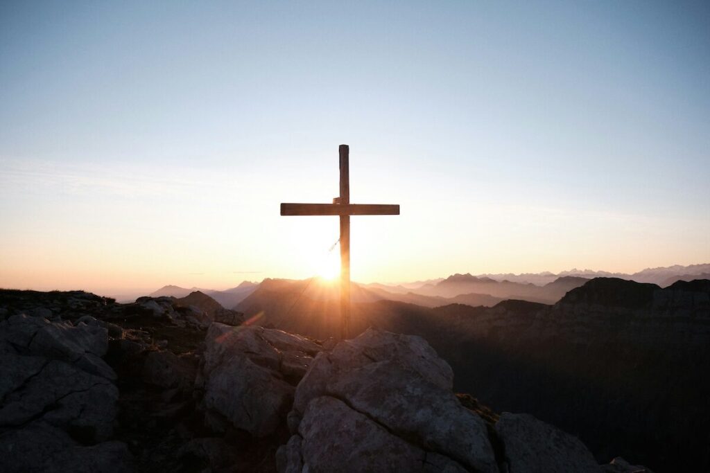A wooden cross stands on a rocky hilltop with the sun rising or setting behind it, casting light over distant mountains.