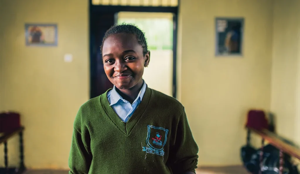 A young student in a green school uniform stands indoors, smiling at the camera. Benches and wall decorations are visible in the background.