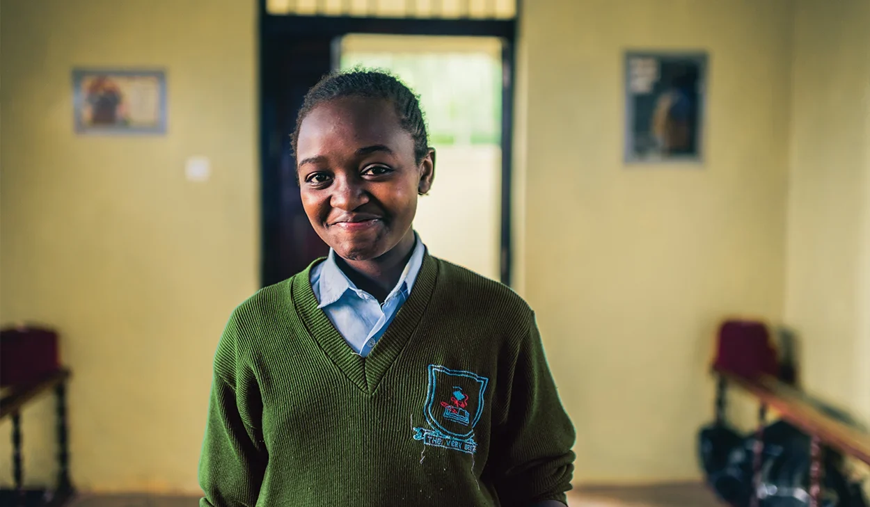 A young student in a green school uniform stands indoors, smiling at the camera. Benches and wall decorations are visible in the background.