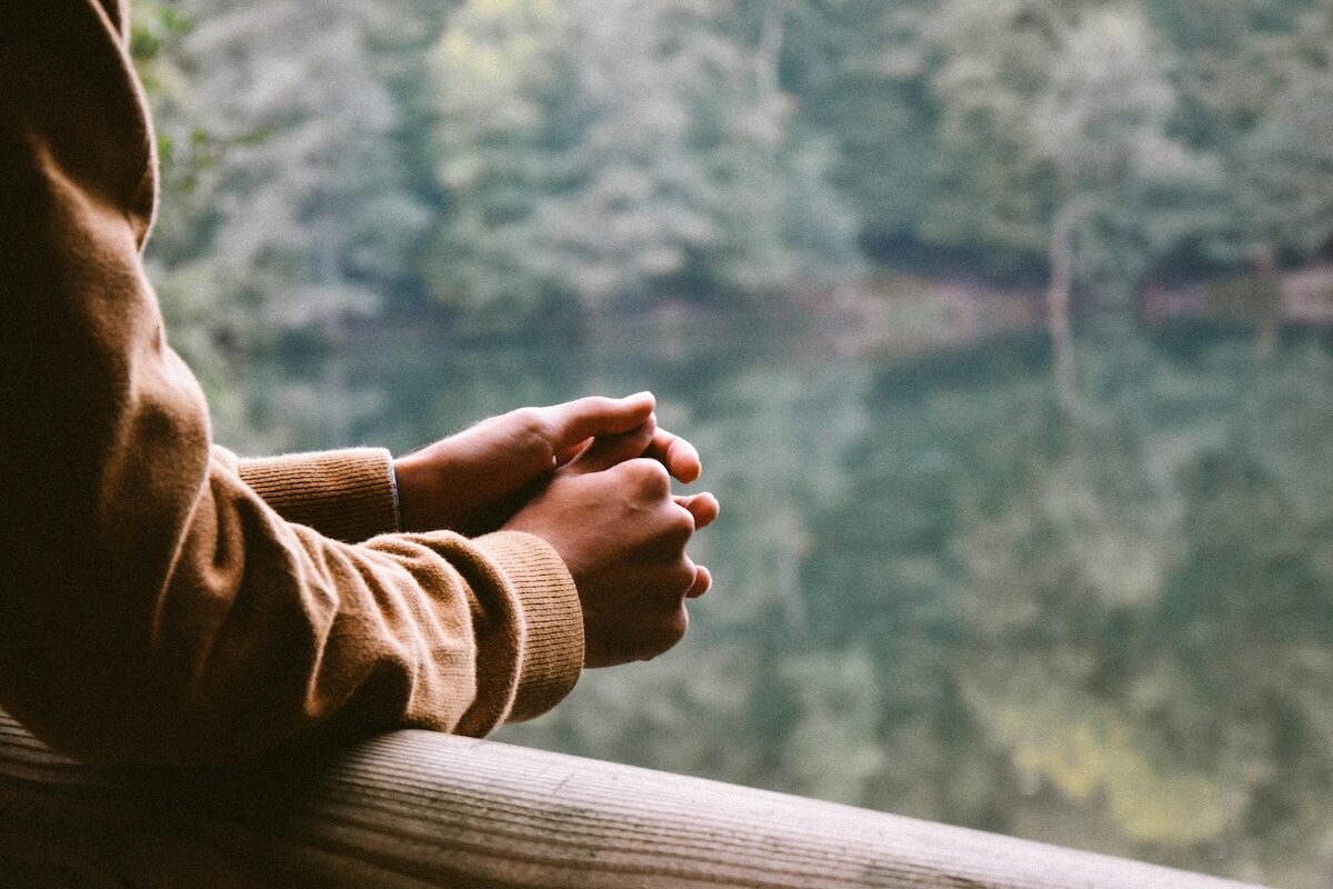 A person in a brown jacket rests their clasped hands on a wooden railing, overlooking a calm body of water with trees in the background.