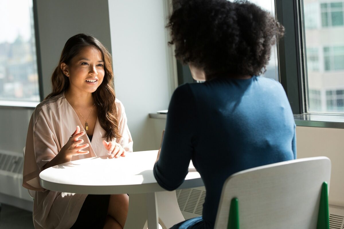Two women sit across from each other at a round table in an office, engaged in a conversation near large windows.