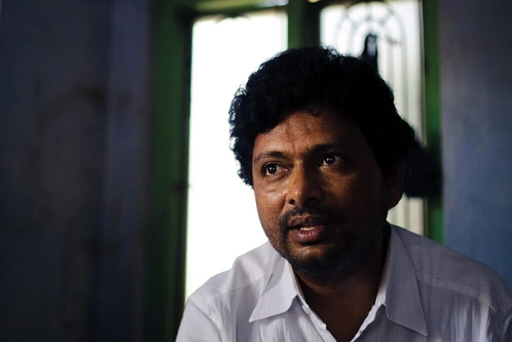 A man with curly hair and a beard, wearing a white shirt, sits indoors in front of a window with green frames and decorative metalwork.