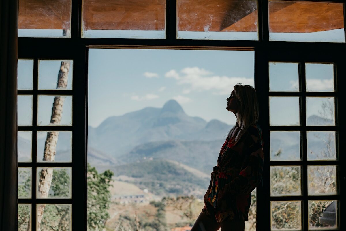 A person stands in silhouette by large windows, looking out at distant mountains on a clear day.
