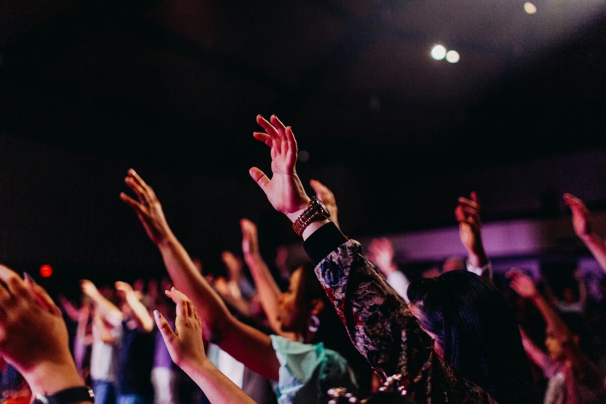 A crowd of people raises their hands in the air during an indoor event, with dim lighting and blurred background.