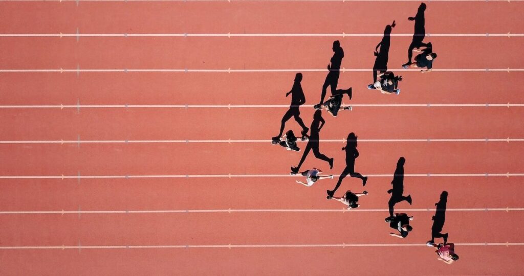 Aerial view of runners racing on a red track, each in separate lanes, casting long shadows across the surface.