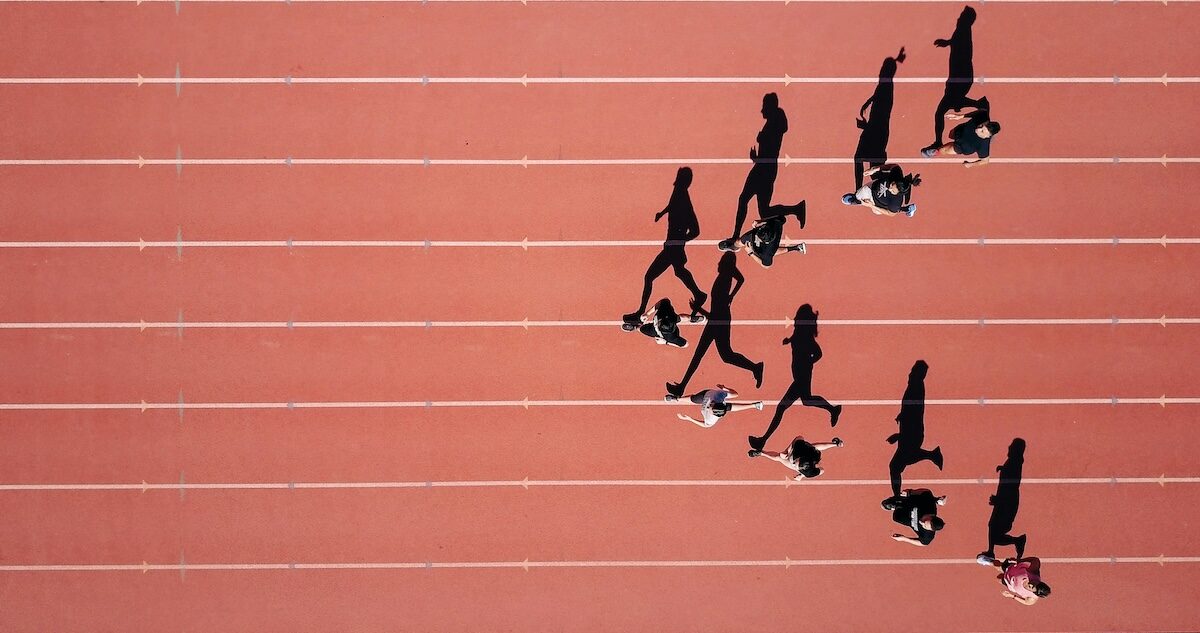 Aerial view of runners racing on a red track, each in separate lanes, casting long shadows across the surface.