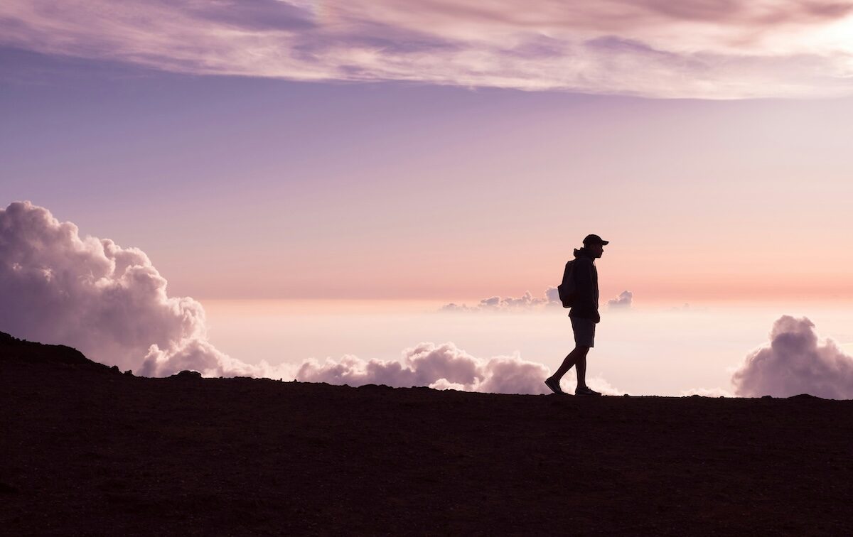 A person wearing a hat and backpack walks along a ridge at sunset, with clouds and a colorful sky in the background.