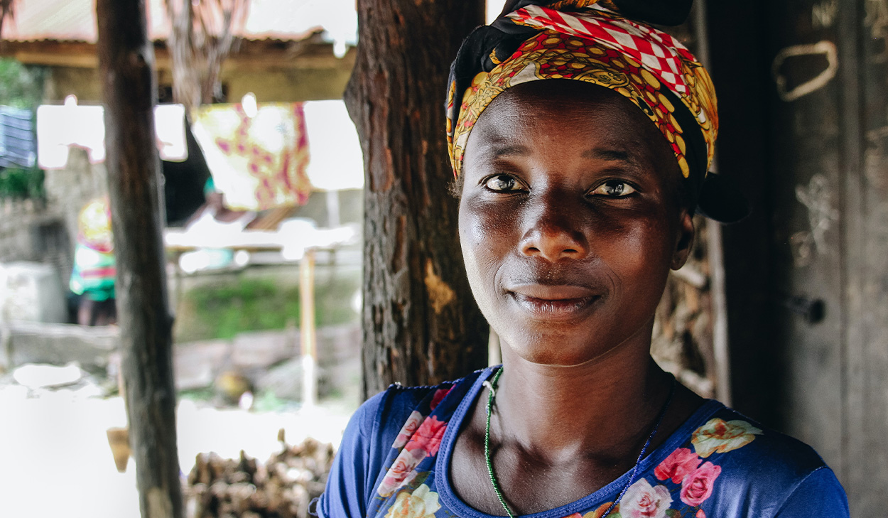 A woman wearing a colorful headscarf and floral shirt stands outside near a wooden structure, looking at the camera.