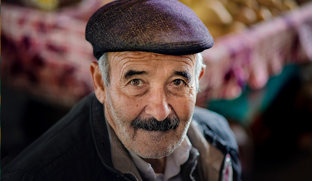 An elderly man with a mustache and flat cap looks at the camera, wearing a jacket and shirt, with a blurred background.
