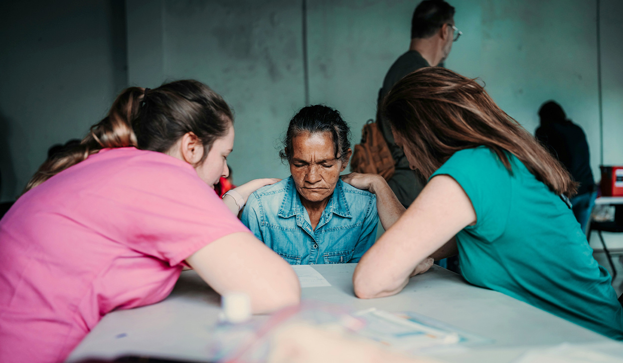 Three women sit at a table, two with heads bowed and hands on the shoulders of the woman in the middle, who appears to be deep in thought.