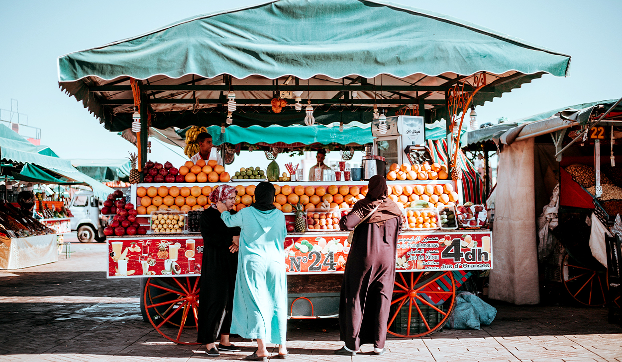 Three people stand at a colorful fruit juice cart under a green canopy at an outdoor market on a sunny day. The cart displays stacked fruit and juice options.