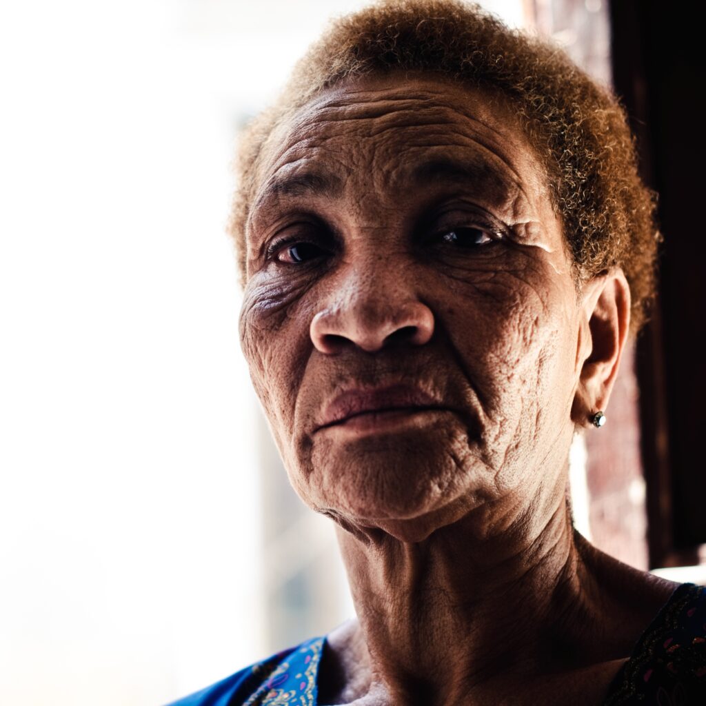 An older woman with short, curly hair and a serious expression stands by a window, partially illuminated by natural light.