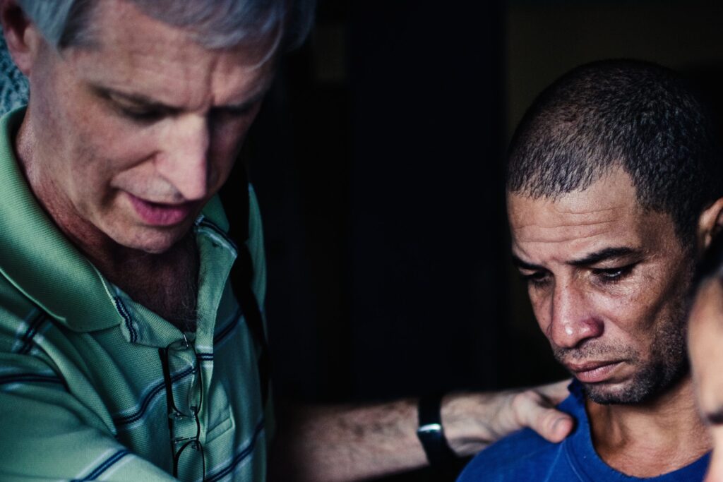 A man in a green striped shirt places his hand on the shoulder of another man who appears sad or distressed, looking downward.