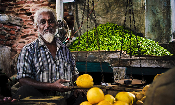 An elderly man with white hair and beard stands at a market stall, holding yellow fruits, with a large pile of green chilies behind him.