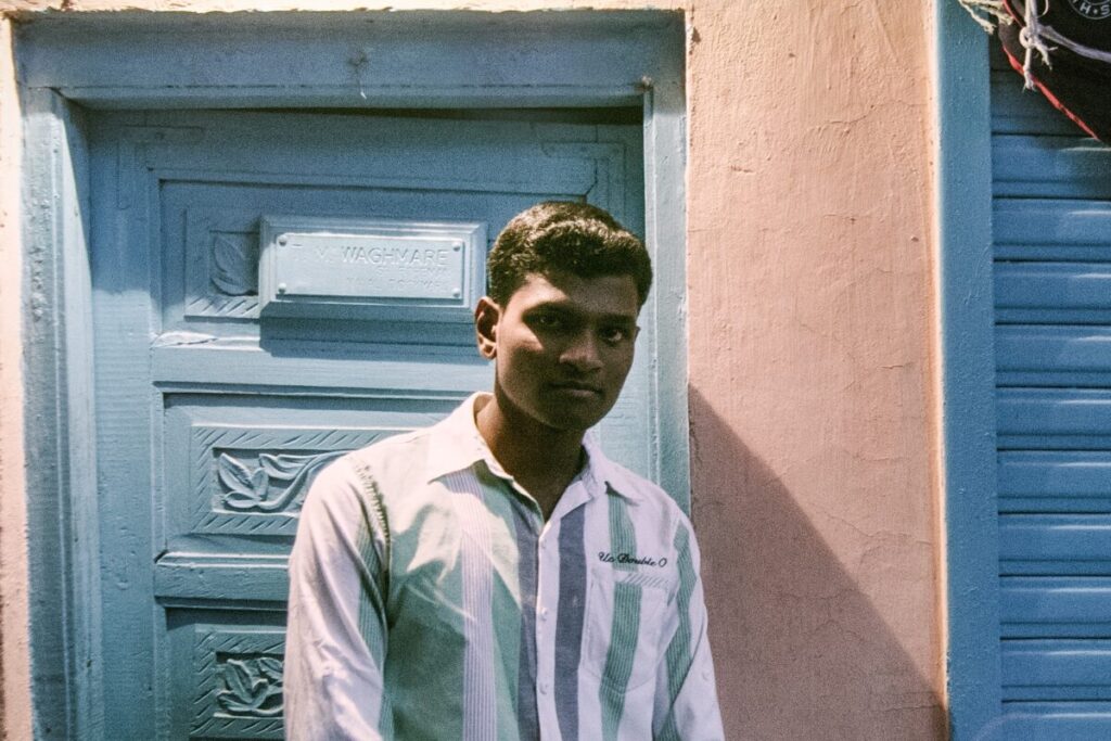 A man in a striped shirt stands in front of a blue door and pink wall, looking slightly toward the camera.