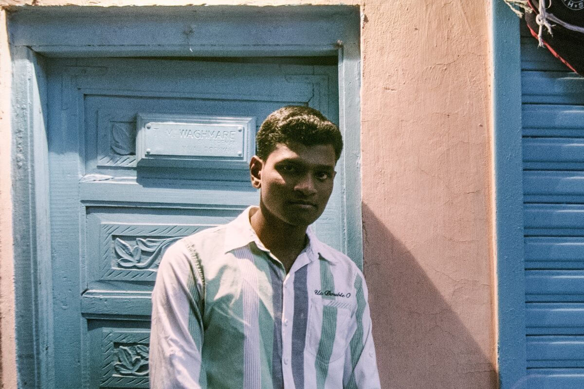 A man in a striped shirt stands in front of a blue door and pink wall, looking slightly toward the camera.