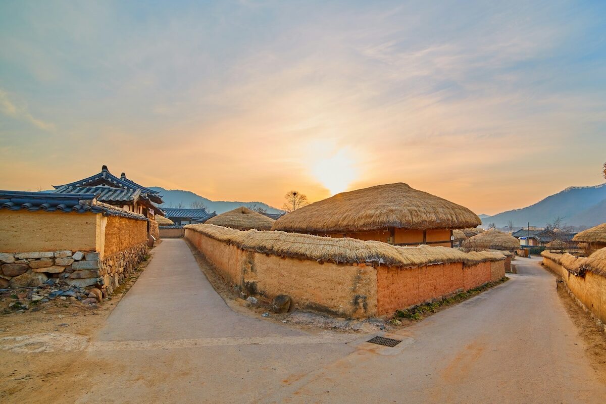 Traditional Korean village with thatched-roof houses and stone walls at sunset, with two paths diverging in the foreground.