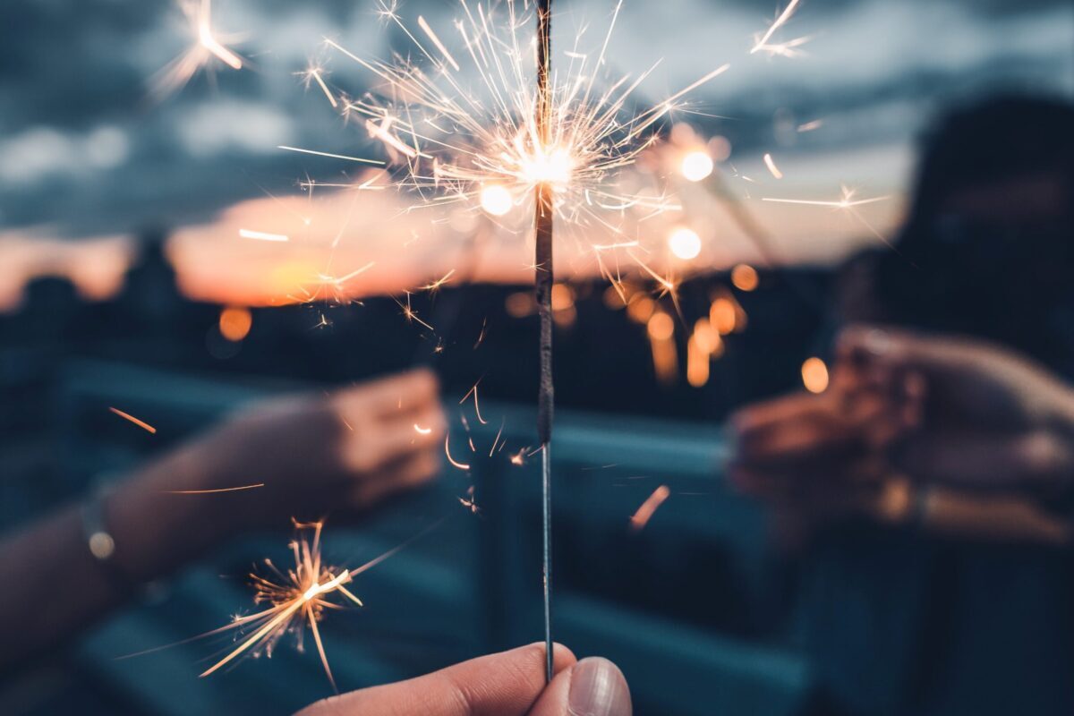 A hand holds a lit sparkler at dusk, with blurred background of people and a sunset sky.