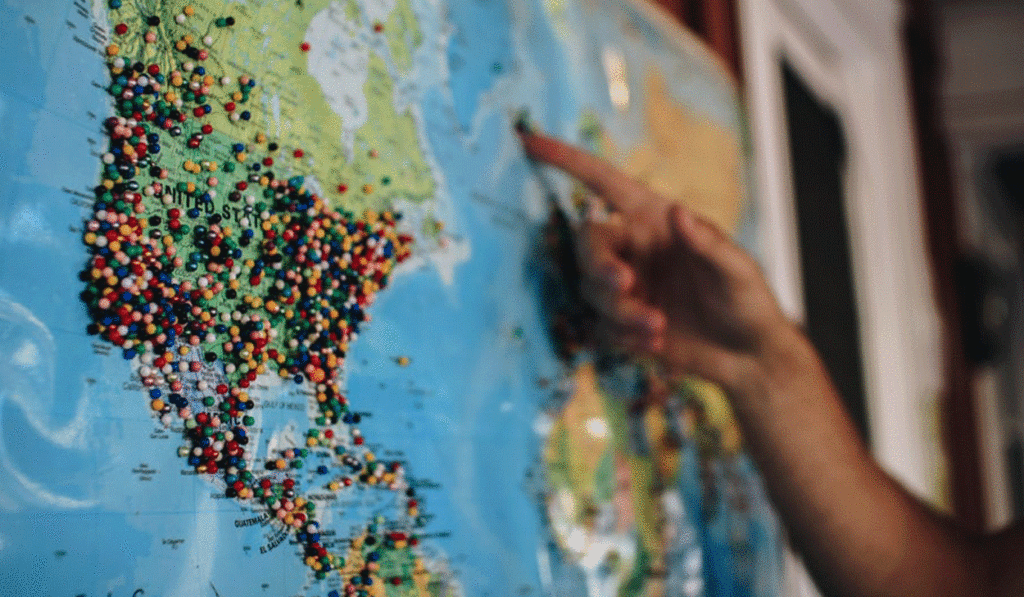 A hand points to a location on a world map covered with numerous colored pins marking various places.