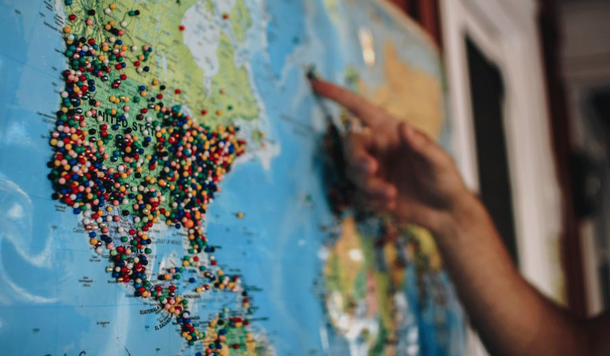 A hand points to a location on a world map covered with numerous colored pins marking various places.