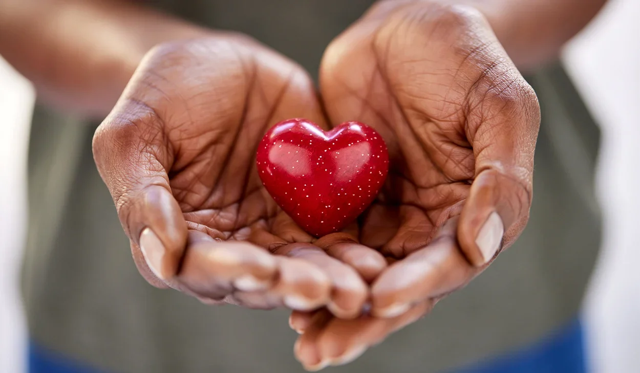 A person holds a small, shiny red heart in both hands, with fingers gently cradling it against a blurred background.