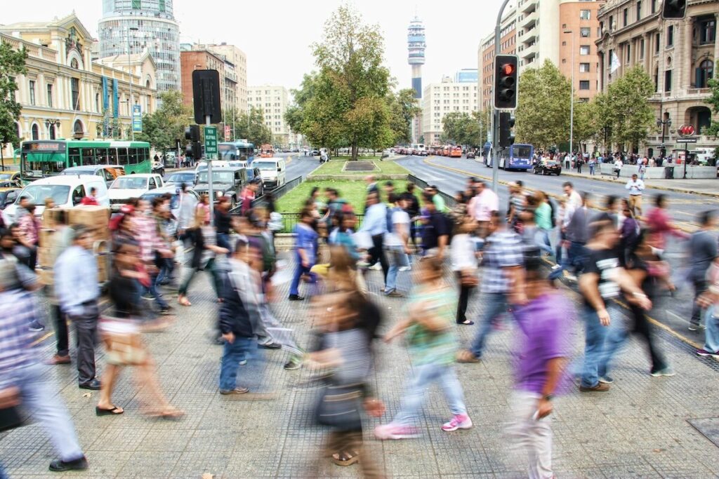 A busy crosswalk in a city with many people walking in both directions; buildings and trees line the street in the background.