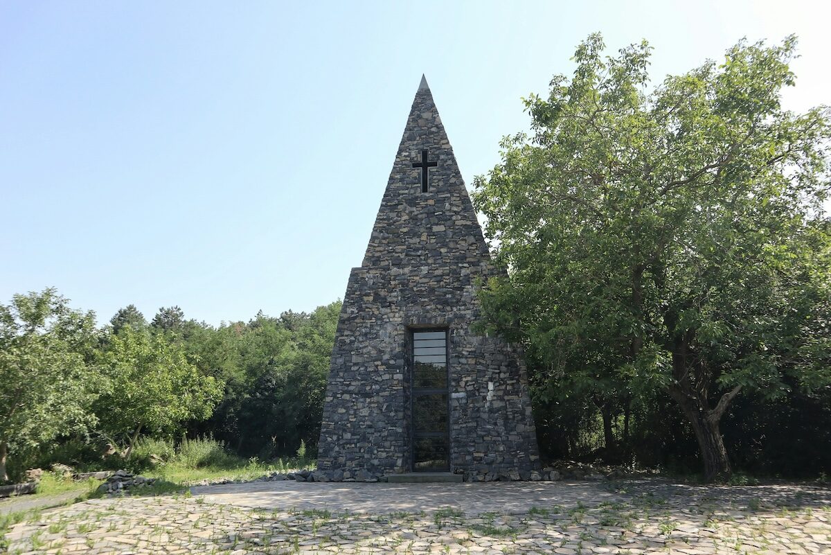 A small stone chapel with a steep triangular roof and a cross above the entrance, surrounded by trees under a clear sky.