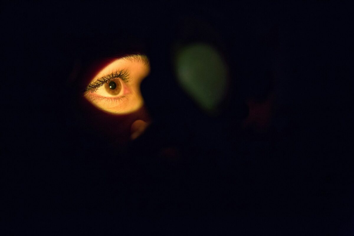 A single human eye illuminated in darkness, surrounded by shadows, with the rest of the face mostly obscured.