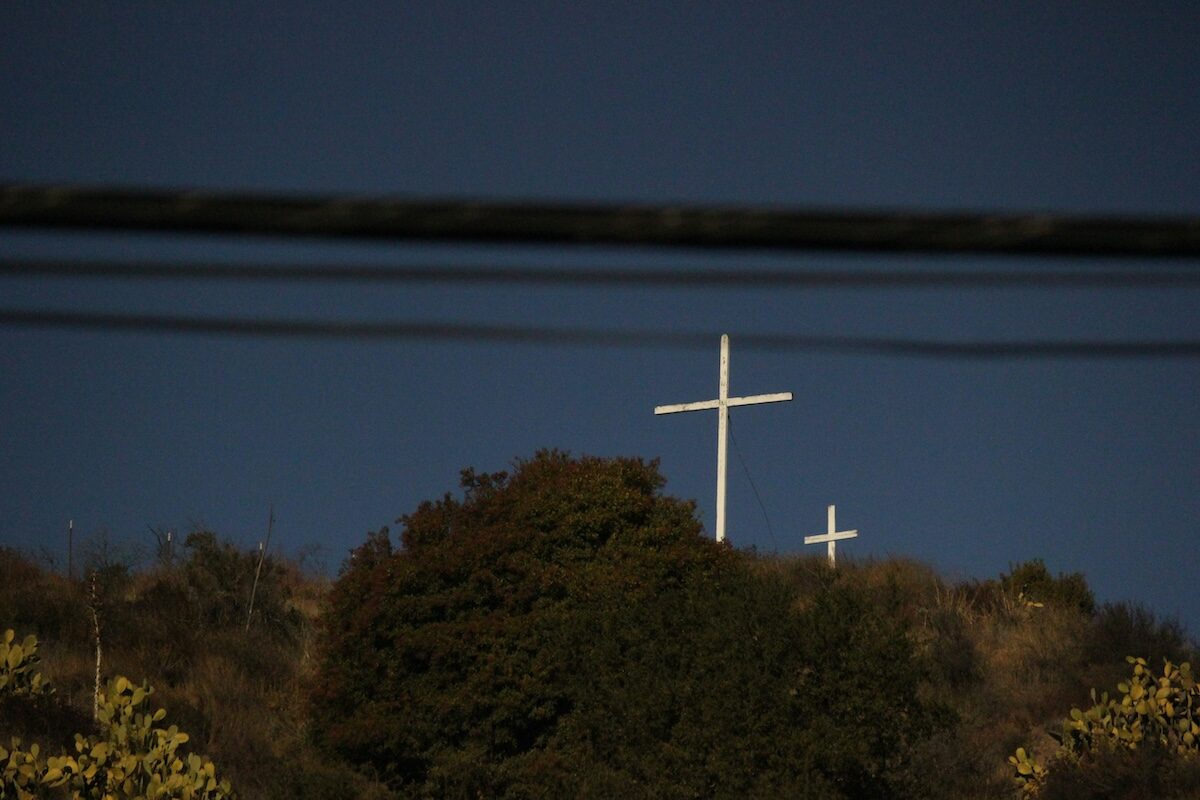 Two white crosses stand on a hilltop against a clear blue sky, with power lines visible in the foreground.
