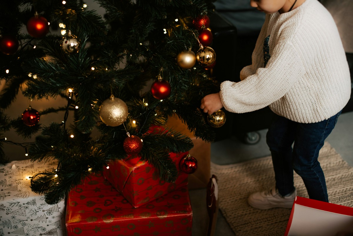 Child decorating a Christmas tree with red and gold ornaments; presents wrapped in red paper are under the tree.